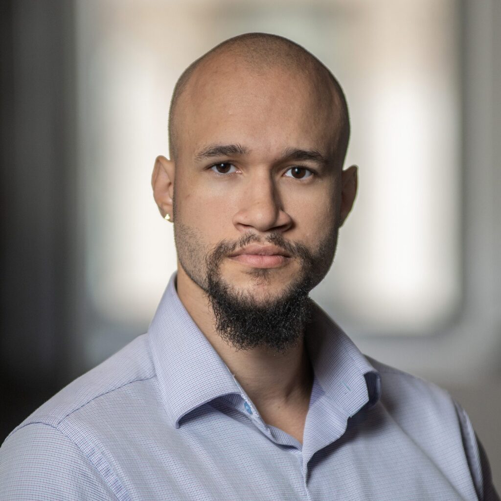 A man with a bald head, chin beard, and mustache, wearing a light blue collared shirt, looks directly at the camera with a neutral expression. The background is softly blurred.