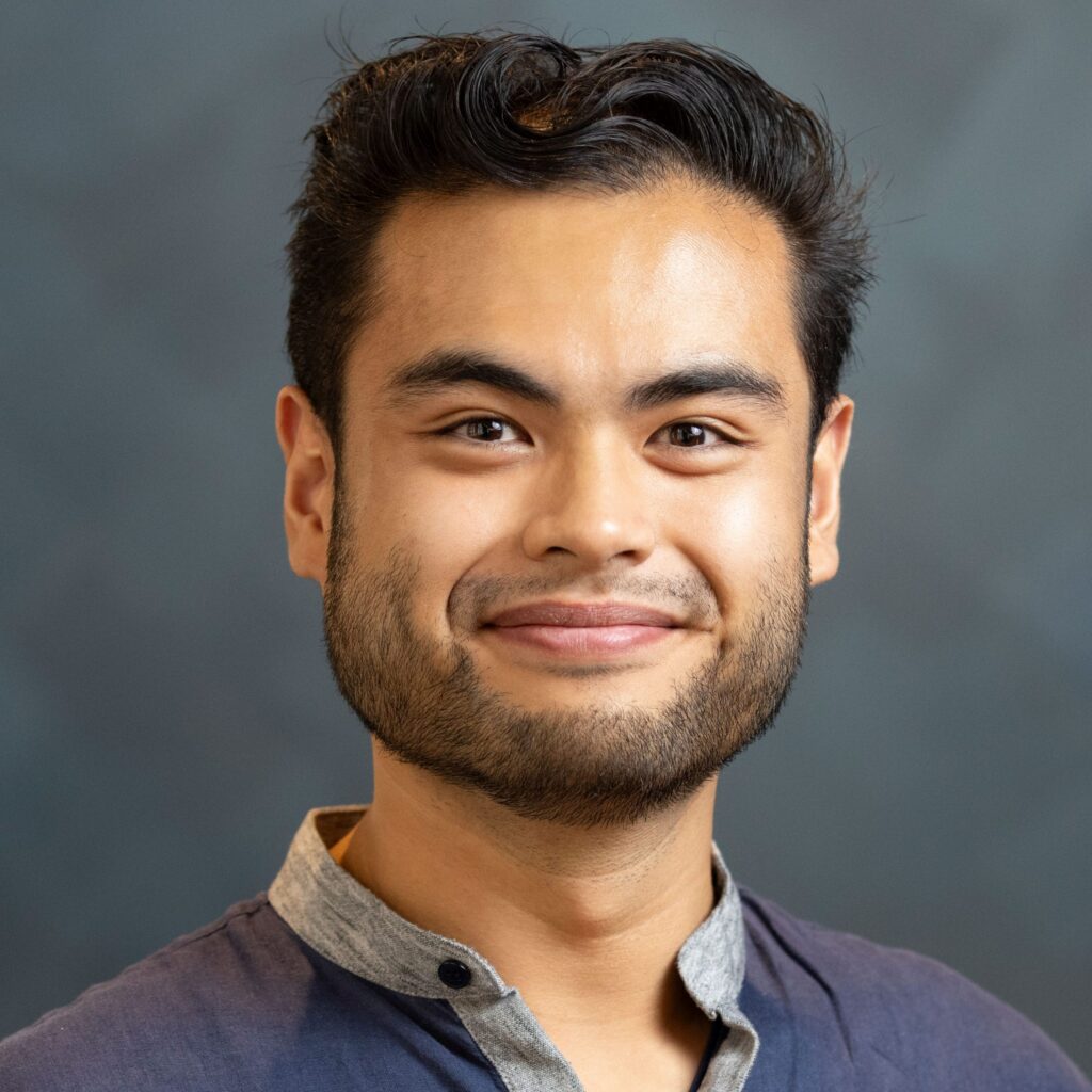 A young man with short black hair and a beard smiles at the camera, wearing a blue shirt with a light gray collar. The background is a soft, blurred gray.