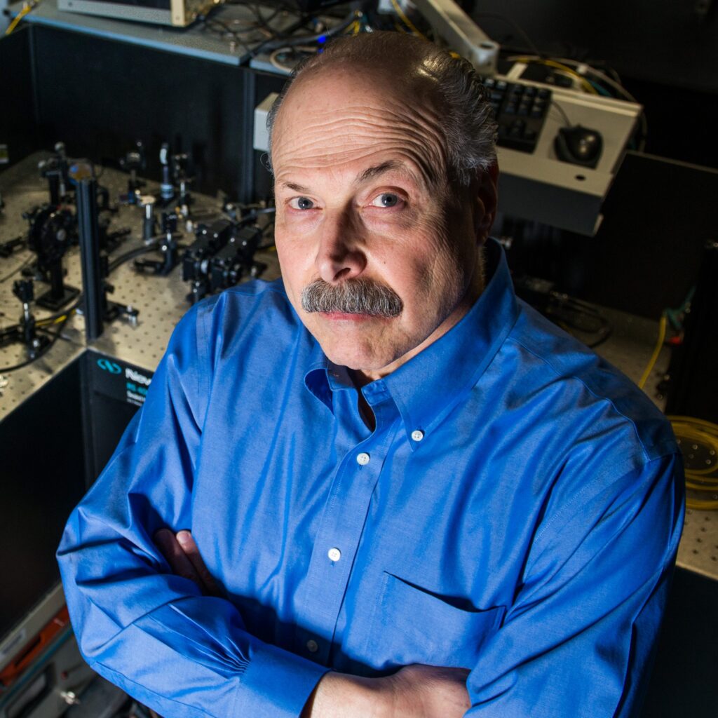 A man with a mustache and blue shirt stands with arms crossed, looking up at the camera. Behind him is scientific equipment on a lab table.