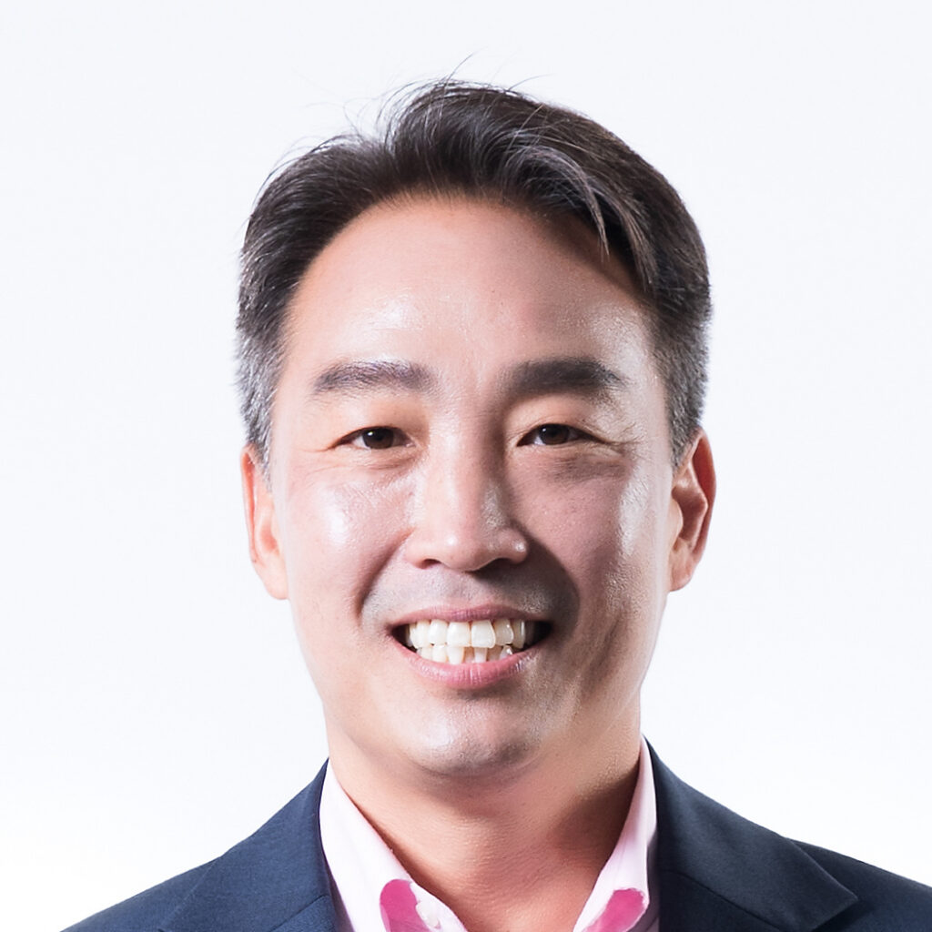 A man with short dark hair smiling, wearing a dark suit jacket and a light pink collared shirt, posing in front of a plain white background.