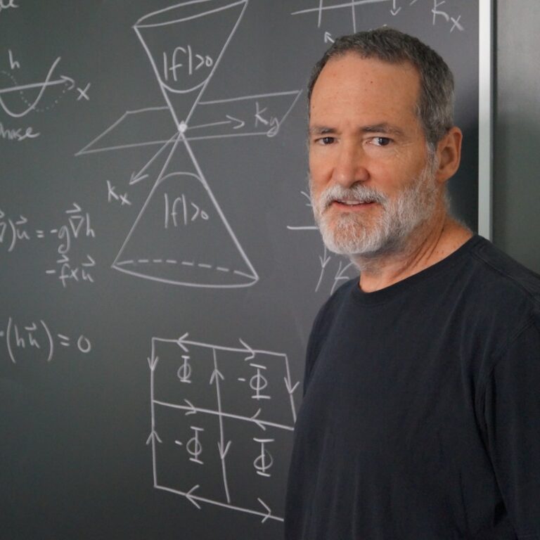 A man with short gray hair and beard stands in front of a chalkboard covered with physics equations and diagrams, wearing a black t-shirt and looking toward the camera.