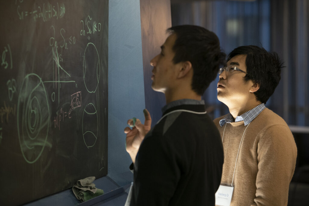 Two people stand by a chalkboard covered in math equations and diagrams. One writes with chalk while the other observes closely, both appearing focused and engaged in problem-solving.