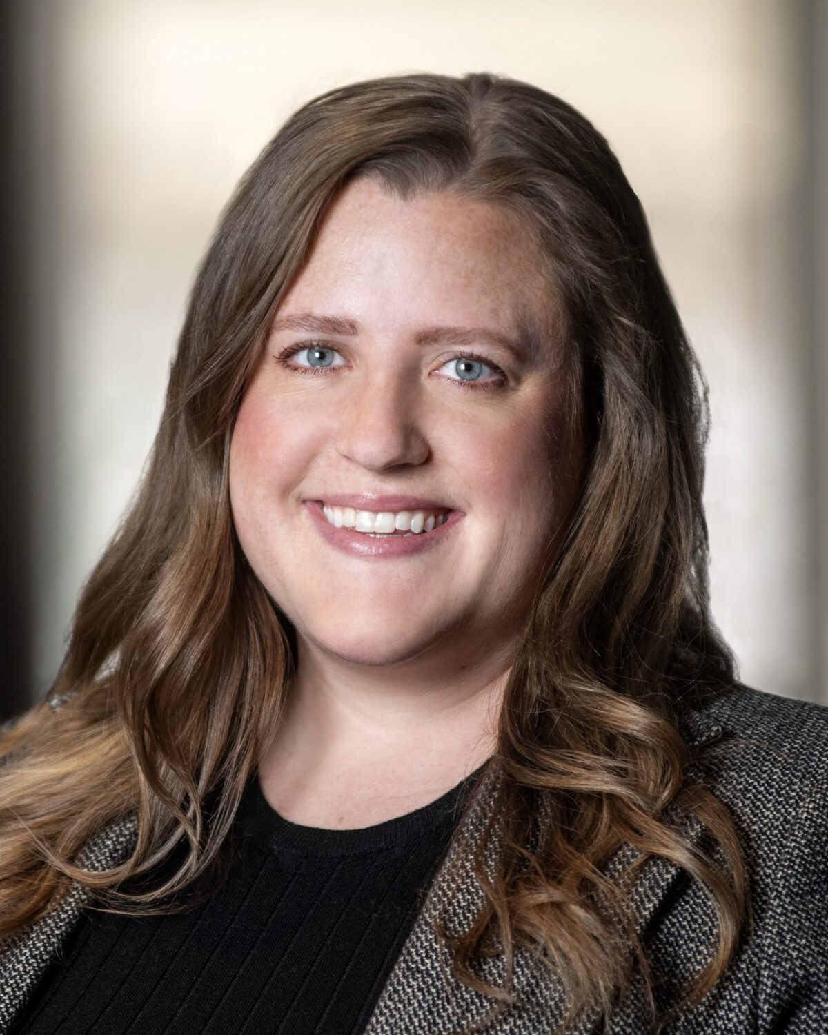 A woman with long, light brown hair smiles at the camera. She is wearing a dark jacket over a black top, and the background is softly blurred in neutral tones.