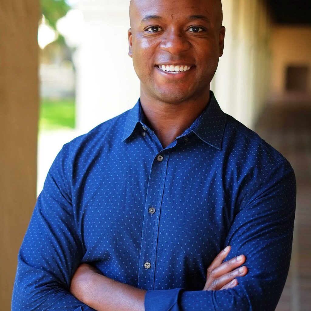 A smiling man with a shaved head wearing a blue, long-sleeve, button-up shirt stands with arms crossed in a sunlit corridor with archways.