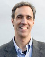 A man with gray hair smiles while wearing a gray suit jacket and light blue dress shirt, standing outdoors against a light, blurred background.