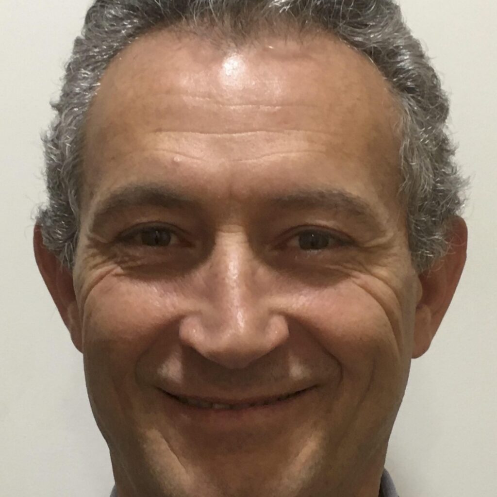 A man with short, curly gray hair is smiling at the camera. He is wearing a dark jacket and a light-colored shirt. The background is plain and light-colored.