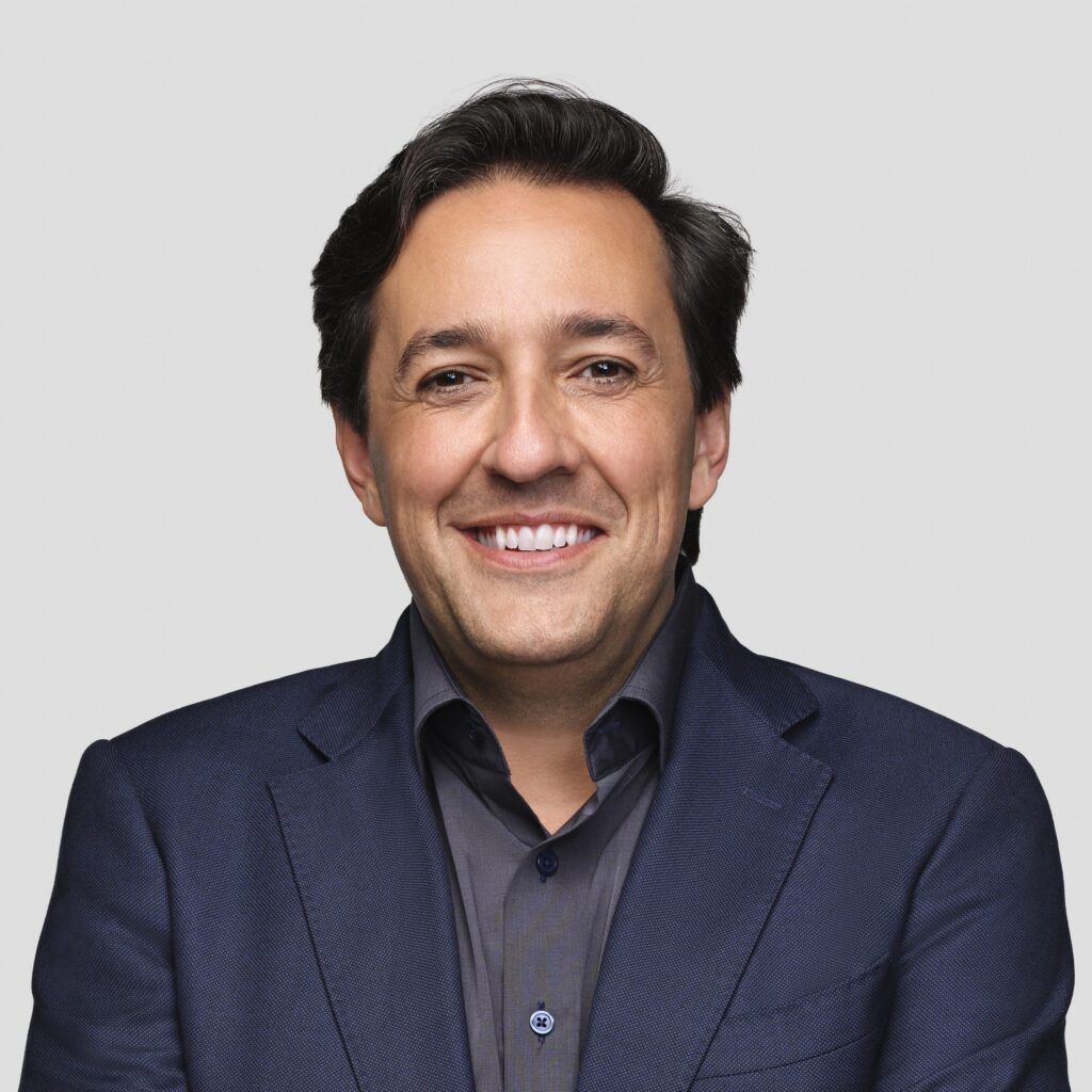A man with dark hair, wearing a navy blazer and dark shirt, smiles at the camera against a plain light gray background.