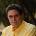 A man wearing glasses and a yellow shirt sits outdoors, looking at the camera. Trees and sunlight are visible in the blurred background.