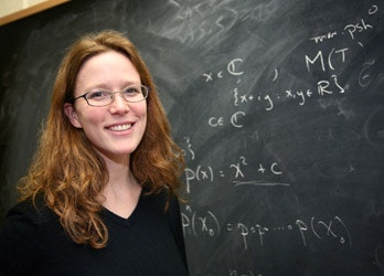 A woman with long red hair and glasses smiles in front of a chalkboard filled with mathematical equations and symbols.