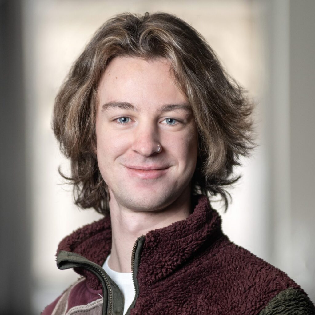 A young person with shoulder-length brown hair and blue eyes smiles while wearing a zipped-up fleece jacket. The background is softly blurred, putting focus on the persons face.
