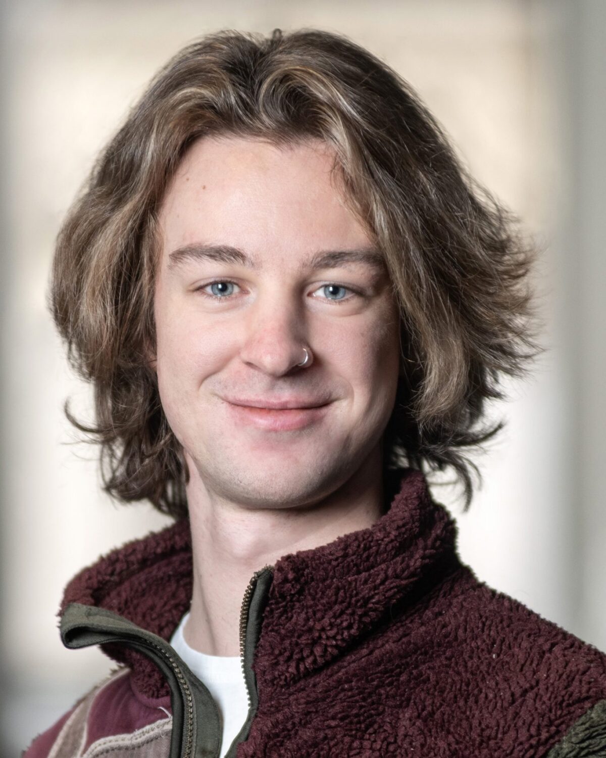 A young person with shoulder-length brown hair and blue eyes smiles while wearing a zipped-up fleece jacket. The background is softly blurred, putting focus on the persons face.