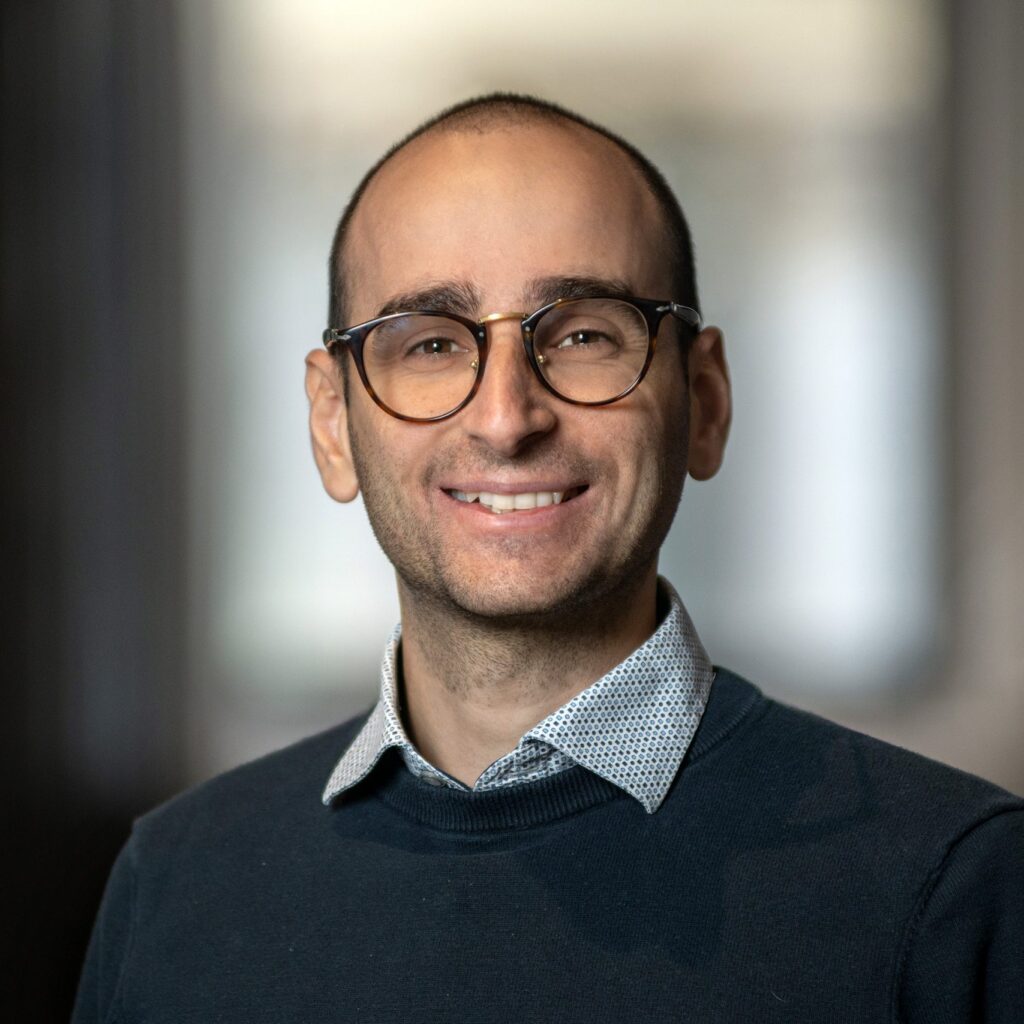 A person with glasses, short dark hair, and a patterned shirt under a dark sweater is smiling at the camera with a blurred background.