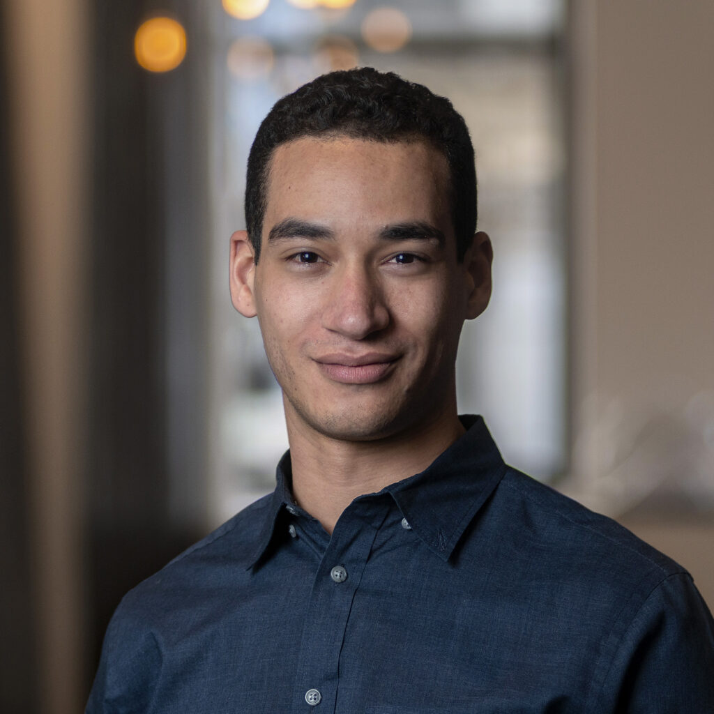 A man with short curly hair and a dark blue shirt smiles softly, standing indoors with blurred windows and warm lights in the background.