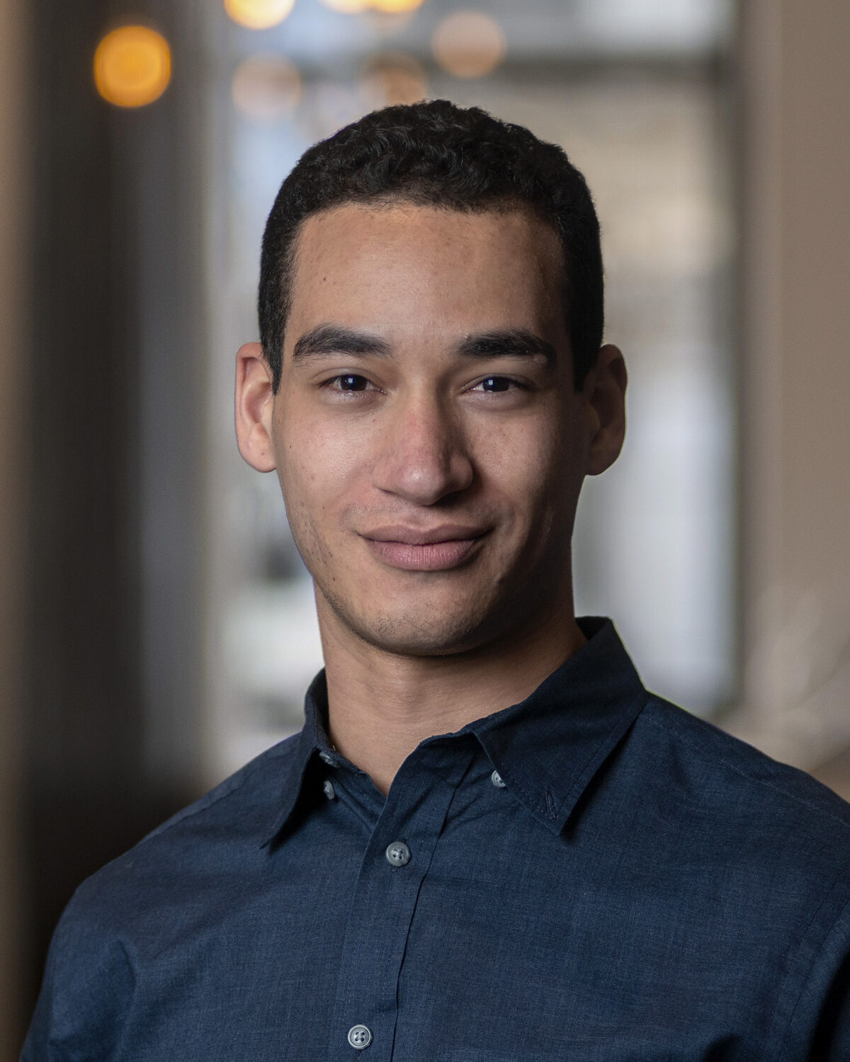 A man with short curly hair and a dark blue shirt smiles softly, standing indoors with blurred windows and warm lights in the background.