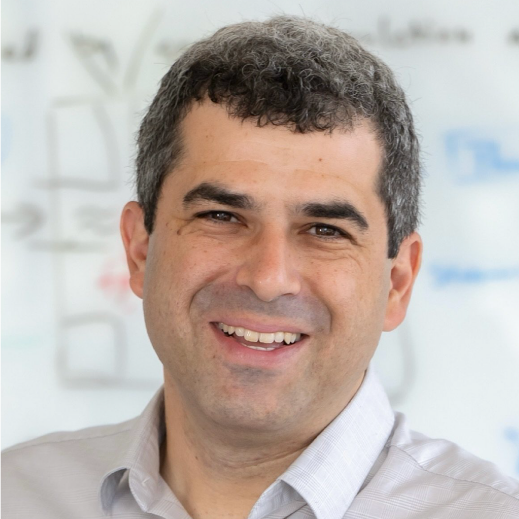 A smiling man with short, curly salt-and-pepper hair wearing a light-colored collared shirt, standing in front of a whiteboard with faint writing and diagrams.