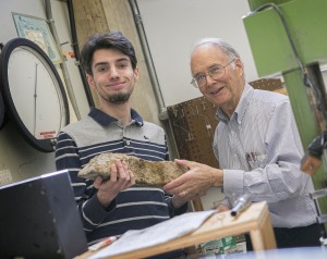 Two men stand indoors and smile at the camera while holding a large rock sample together, surrounded by scientific equipment and a large clock on the wall.