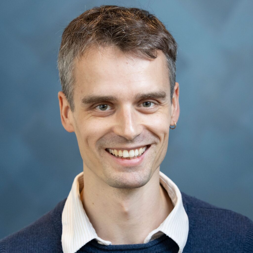 A smiling man with short brown hair wearing a blue sweater over a white collared shirt stands in front of a blue blurred background.