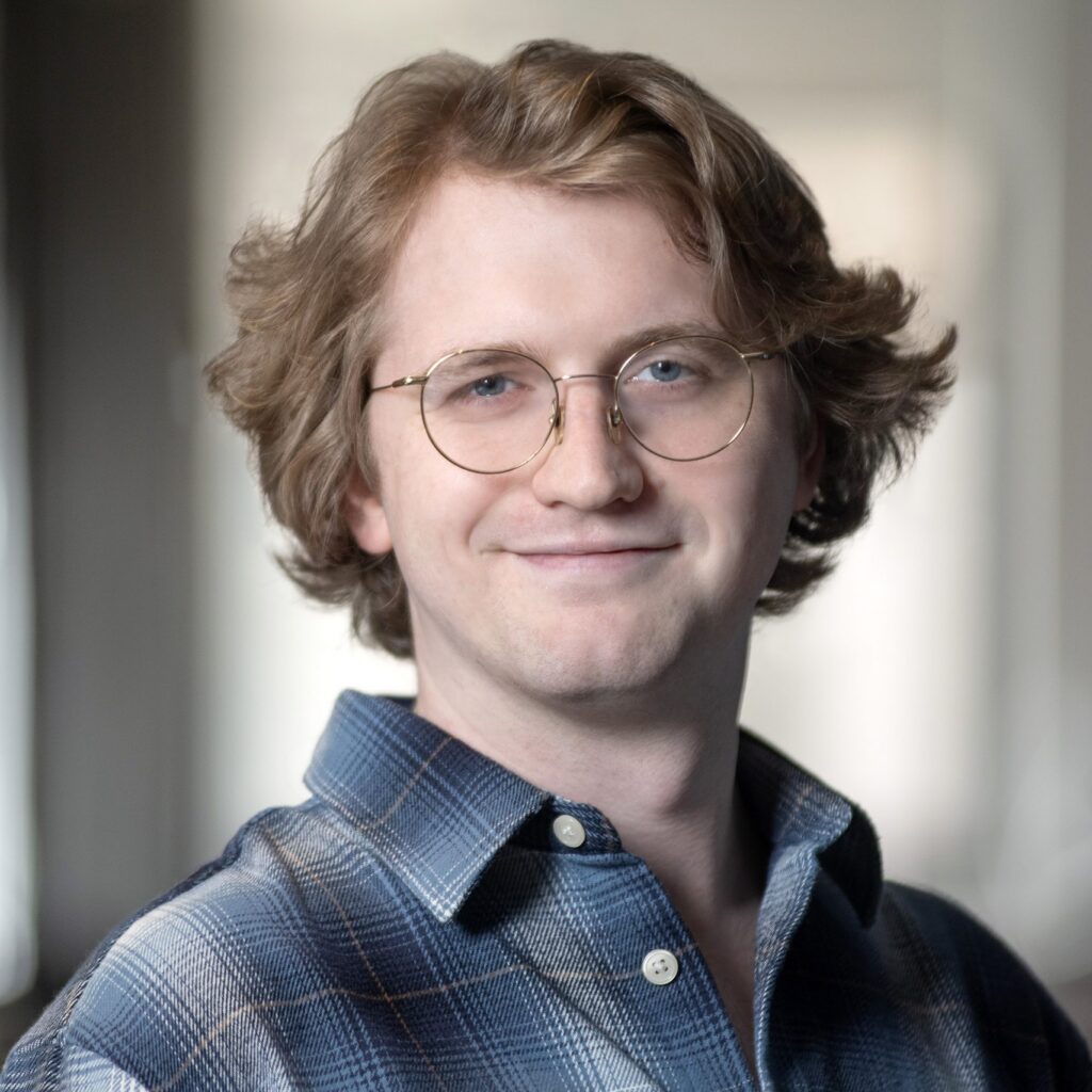 A young person with wavy light brown hair and glasses, wearing a blue plaid shirt, smiles softly while standing indoors with a blurred background.