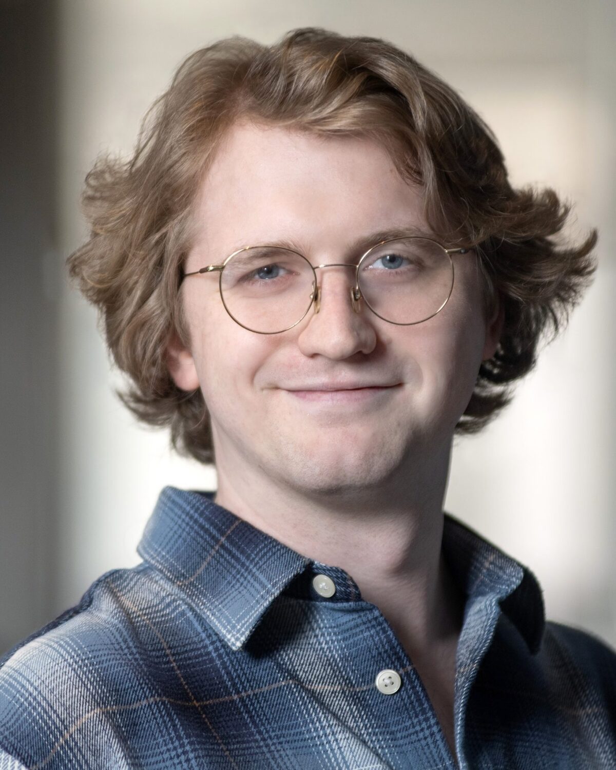 A young person with wavy light brown hair and glasses, wearing a blue plaid shirt, smiles softly while standing indoors with a blurred background.