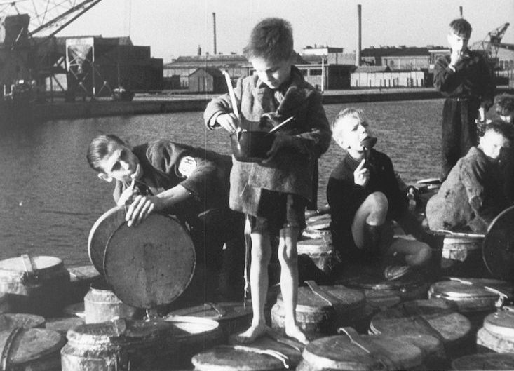 A group of children, some eating, gather near a dock with large metal containers. One boy stands barefoot on the containers, holding and stirring a pot. Industrial buildings and cranes are in the background.