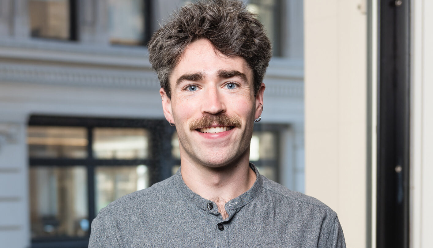 A person with grayish hair and a mustache is smiling indoors, wearing a gray button-up shirt. There is a large window behind them showing the exterior of a building.