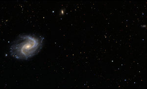 A spiral galaxy with bright arms is seen on the left side of the image, surrounded by stars and distant galaxies scattered across a dark, star-filled space background.