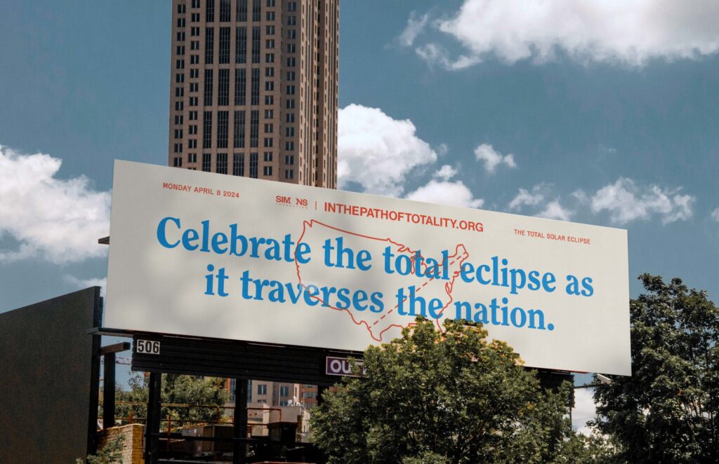 A billboard reads, Celebrate the total eclipse as it traverses the nation, with a map of the U.S. and info about the April 8, 2024 total solar eclipse. Trees and a tall building are in the background.
