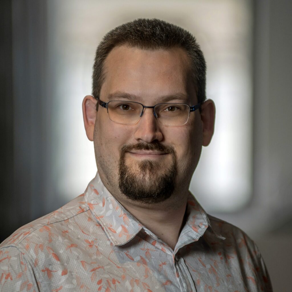 Man with short hair, glasses, a mustache, and goatee smiles softly. He wears a light-colored, patterned shirt and stands indoors with a blurred background.
