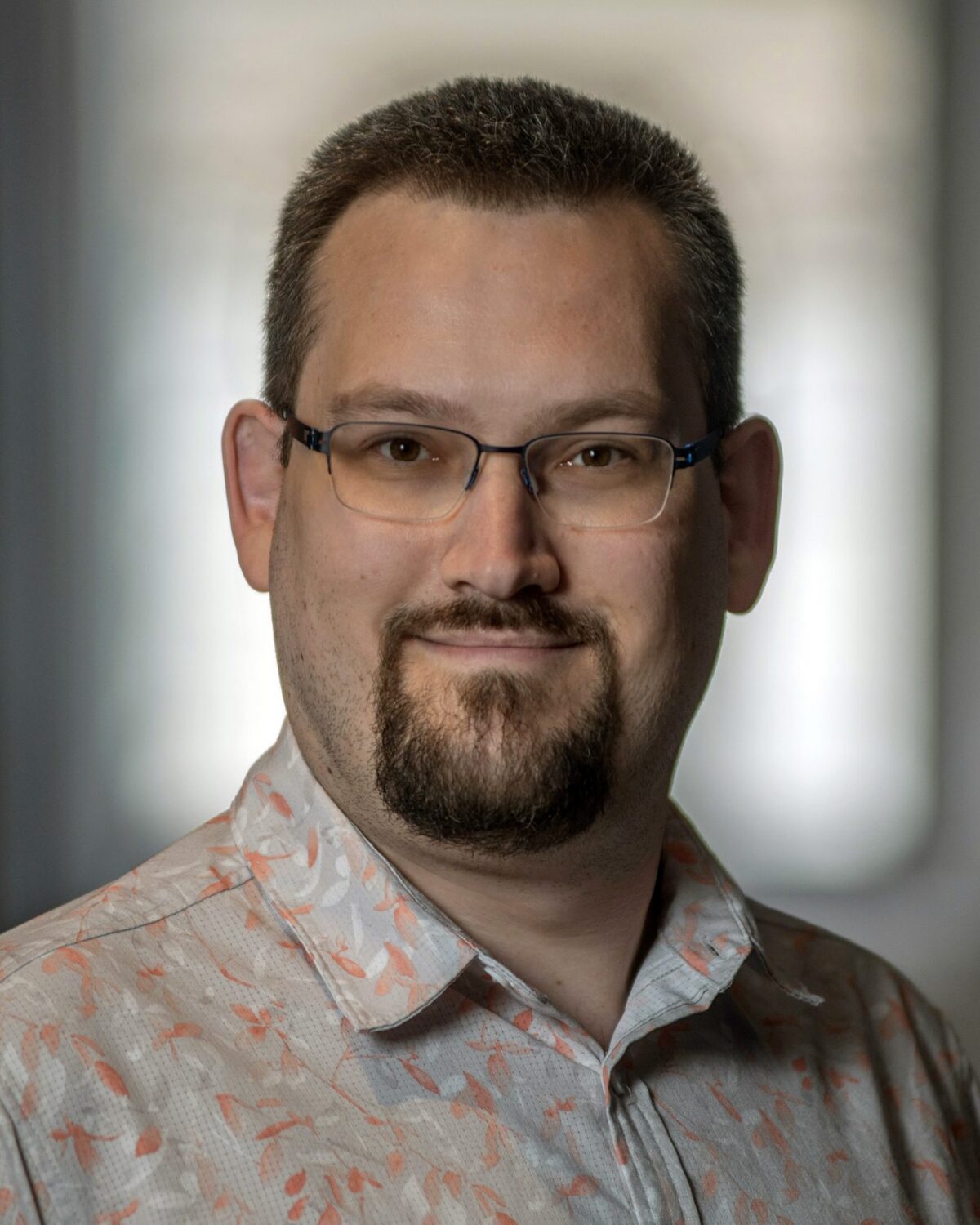 Man with short hair, glasses, a mustache, and goatee smiles softly. He wears a light-colored, patterned shirt and stands indoors with a blurred background.