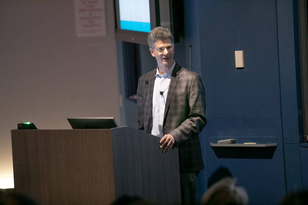 A man in a suit jacket stands at a podium, speaking to an audience in a lecture hall. A microphone is clipped to his jacket, and a screen is visible behind him.