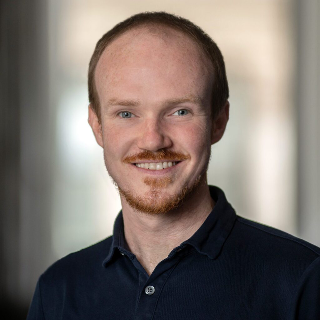 A man with short reddish hair and a beard, smiling, wears a dark polo shirt. The background is softly blurred, bringing focus to his face.
