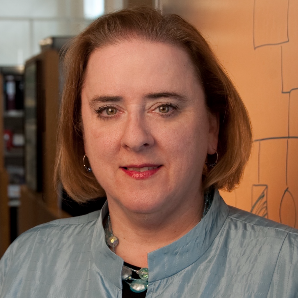 A woman with short light brown hair, wearing a blue jacket and necklace, stands indoors in front of an orange wall with diagrams.