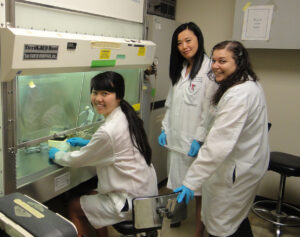 Three women in lab coats and blue gloves work in a laboratory. One is seated at a biosafety cabinet handling equipment, while the other two stand nearby, smiling at the camera.