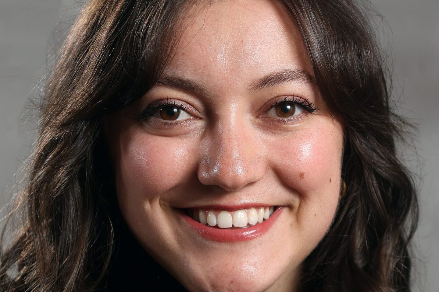 A woman with long wavy brown hair and a black top smiles at the camera. The background is a neutral gray.