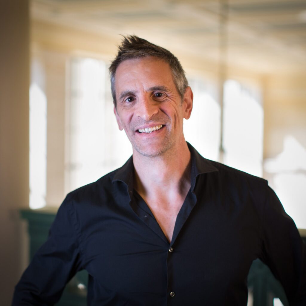 A smiling man with short hair wearing a black shirt stands indoors in soft, natural light with a blurred background.