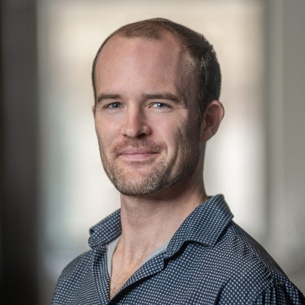 A man with short hair and a beard, wearing a dark patterned shirt, smiles slightly while looking at the camera. The background is softly blurred.