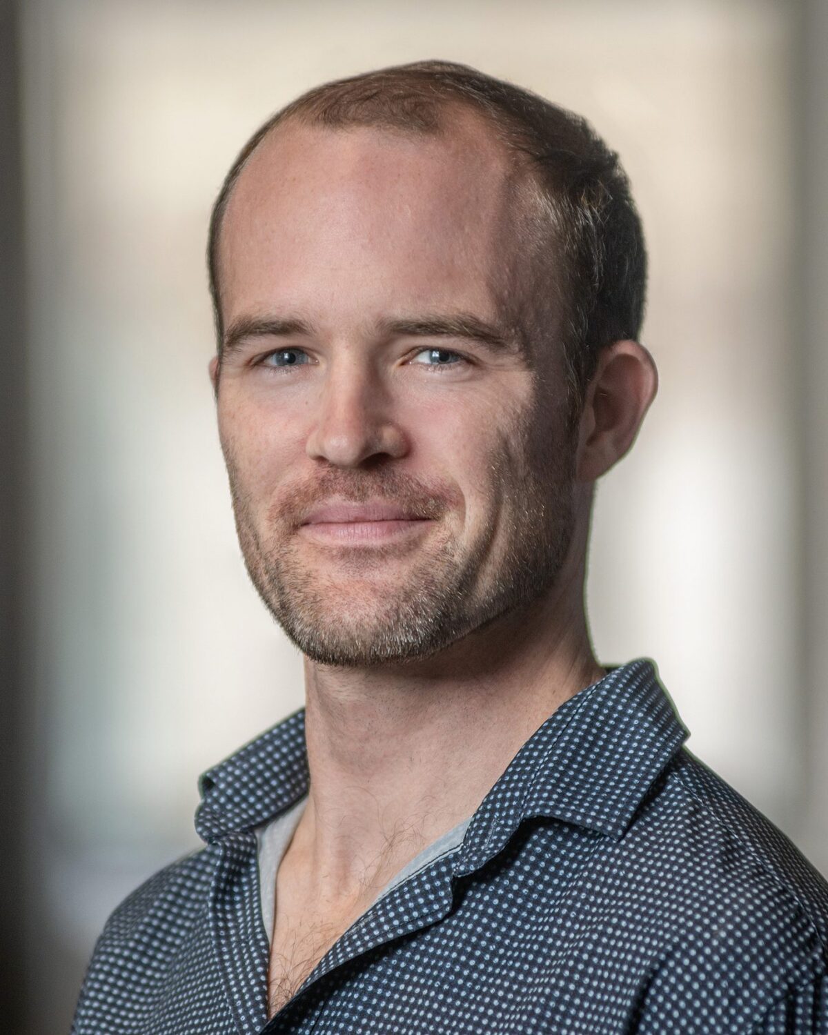A man with short hair and a beard, wearing a dark patterned shirt, smiles slightly while looking at the camera. The background is softly blurred.