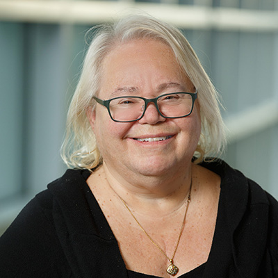 Smiling older woman with long white hair and glasses, wearing a black top and gold necklace, standing in front of a blurred background with large windows.