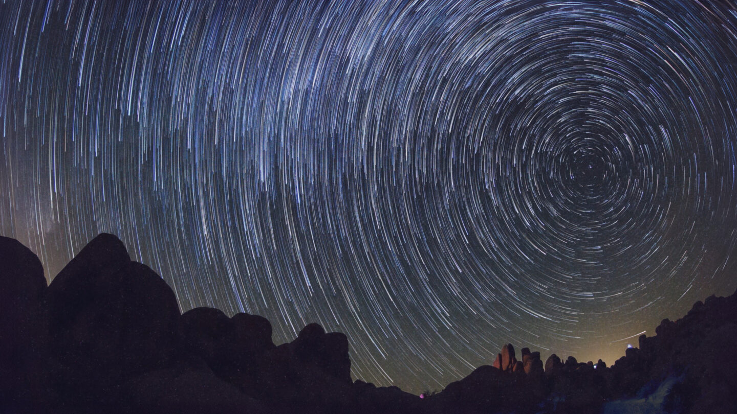 Long exposure photo of a night sky showing star trails in circular patterns above dark, rocky silhouettes, creating a sense of motion and highlighting the rotation of the Earth.