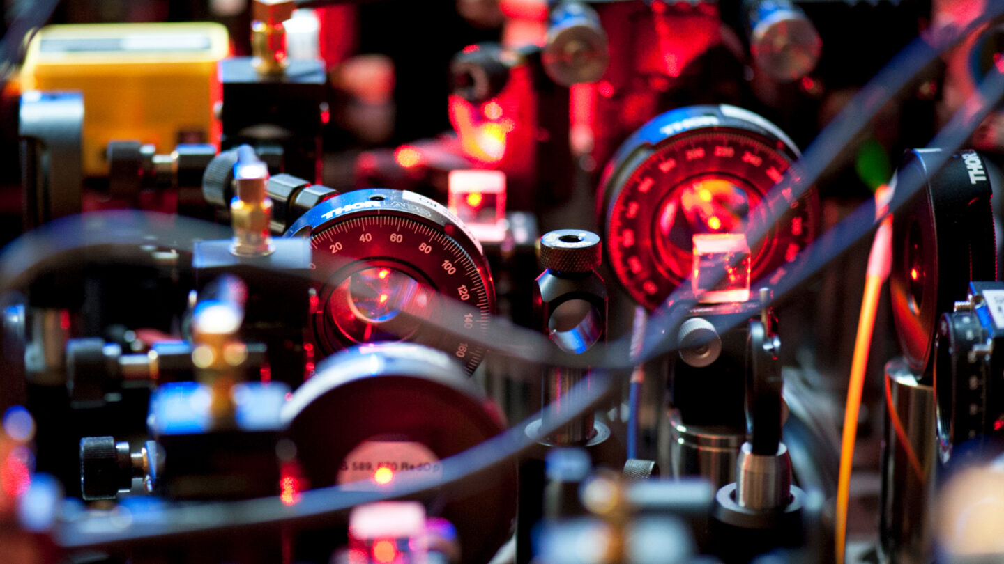 Close-up view of a complex scientific experiment setup with various optical instruments, lenses, dials, and wires, illuminated by red and orange lights, possibly used for physics or laser research.