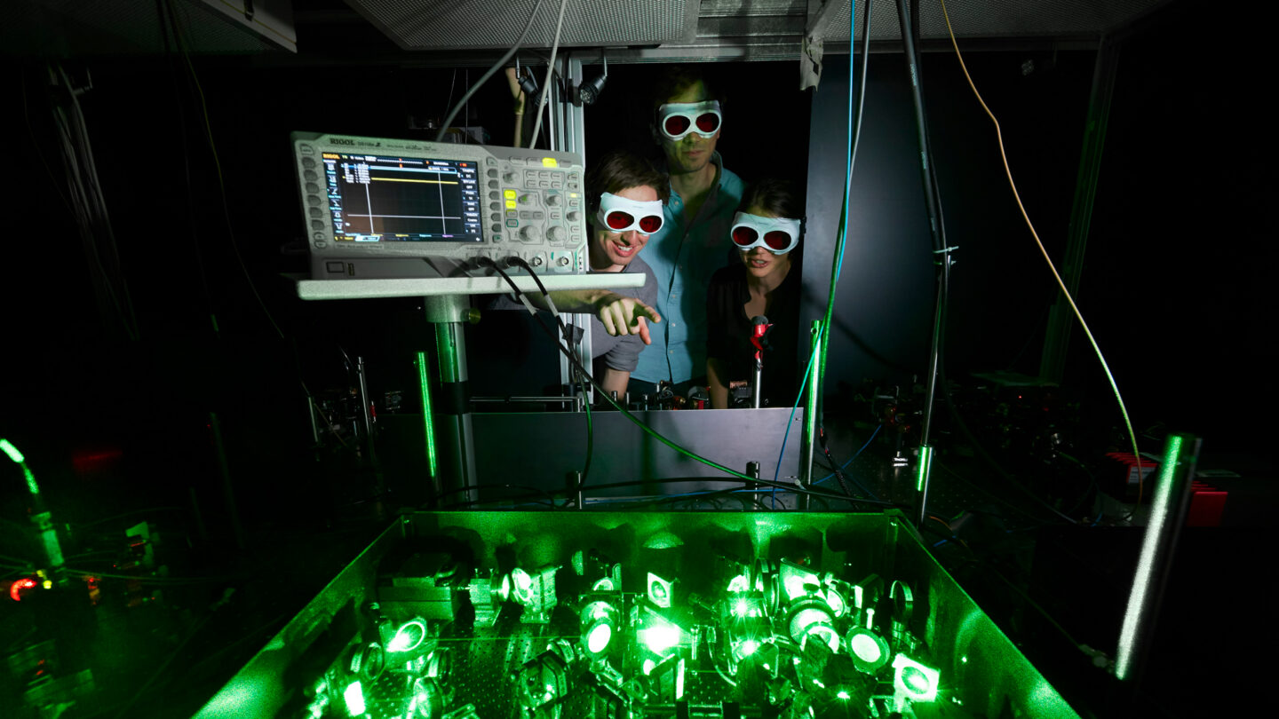 Photo of glowing green tabletop laser equipment with researchers looking on.