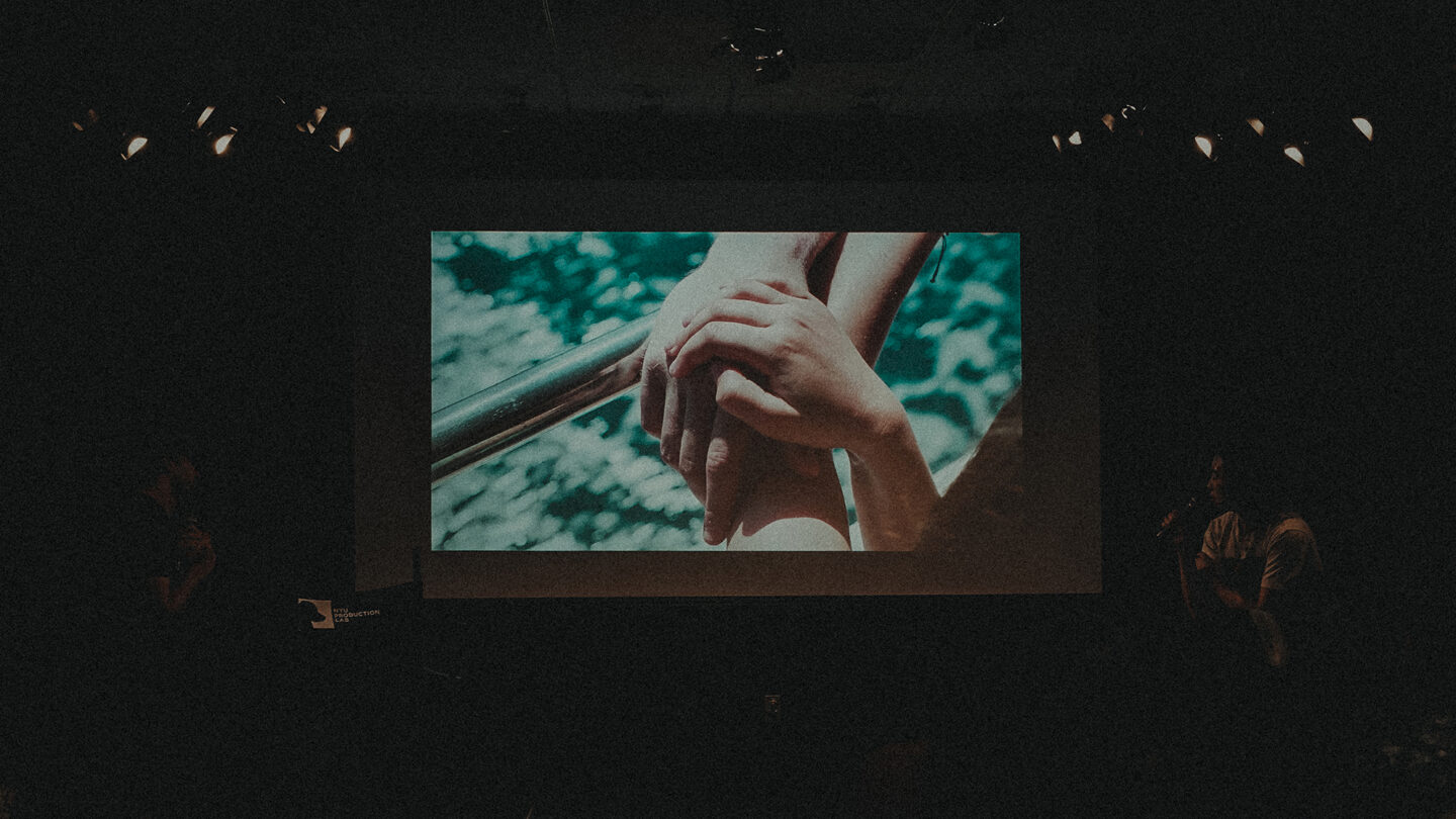 A photo taken at the screening of 'Symbiosis,' showing a dark auditorium with a film still of two clasped hands on the screen.