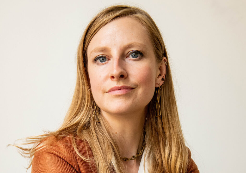 A woman with long blonde hair, wearing a brown blazer, gold earrings, and a gold necklace, looks confidently at the camera against a plain light background.