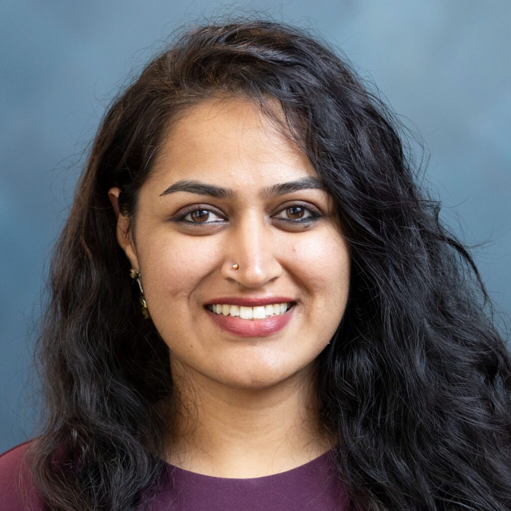 A woman with long wavy dark hair, brown eyes, and a nose stud smiles at the camera. She is wearing a maroon top and gold earrings, with a blue-gray background behind her.