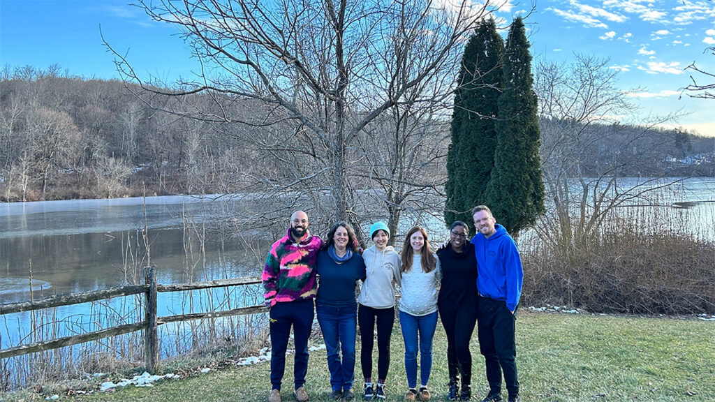 Six people standing outdoors in front of a lake.