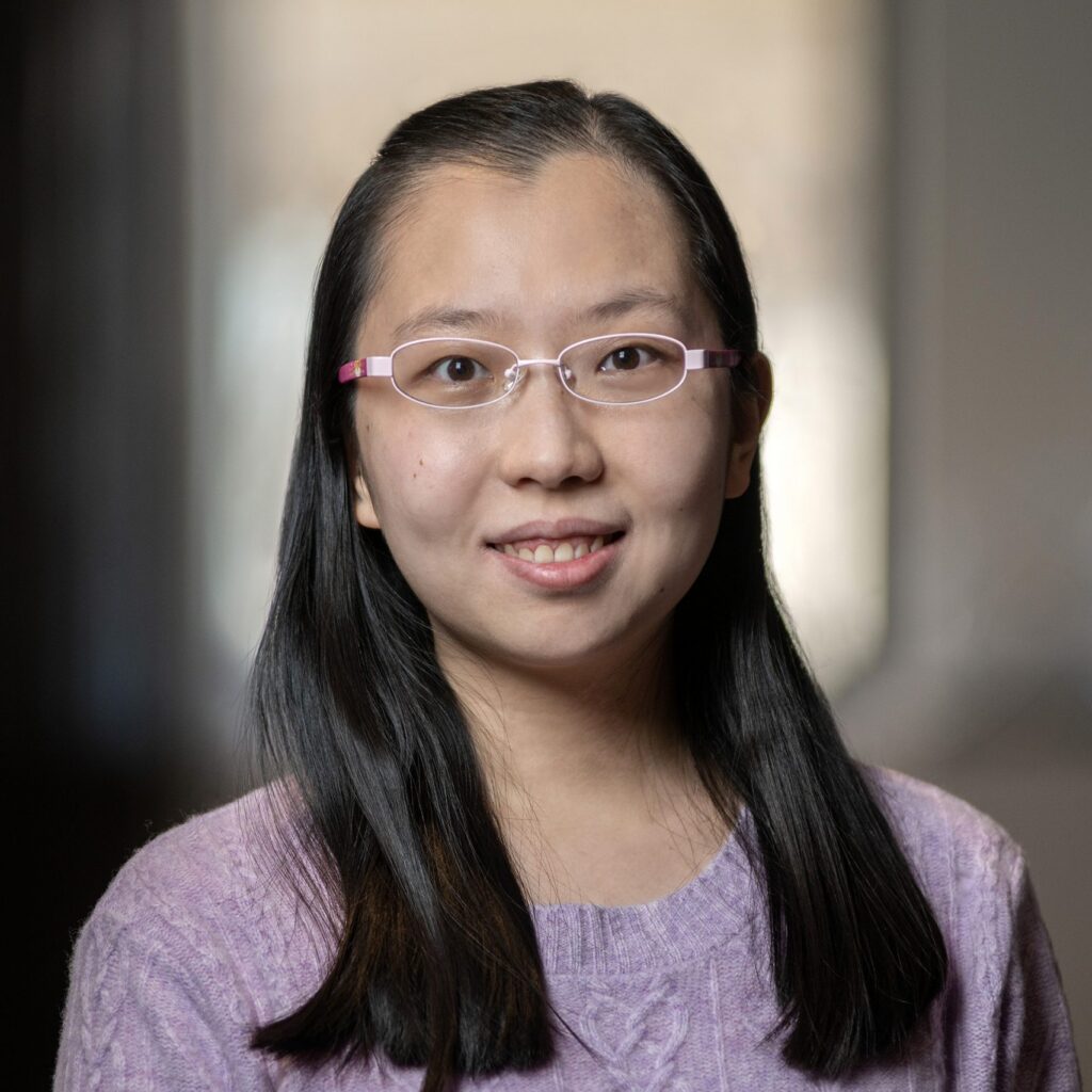 A woman with long dark hair and glasses smiles at the camera. She is wearing a light purple sweater and is standing indoors with a blurred background.