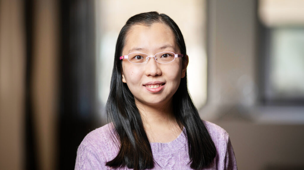 A woman with long black hair, glasses, and a light purple sweater smiles at the camera. The background is softly blurred with neutral tones.