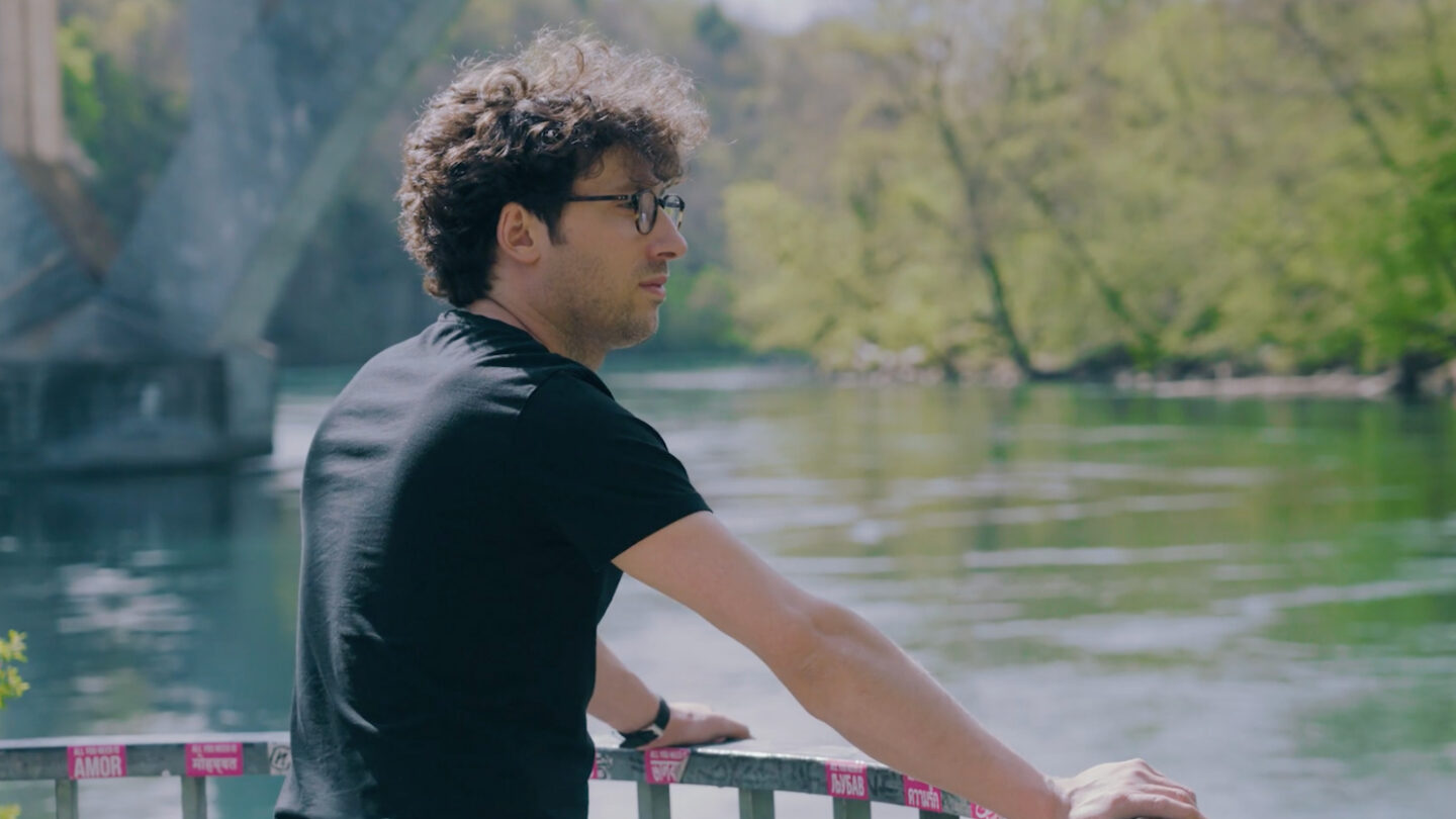 A person with curly hair and glasses stands by a river, leaning on a railing, looking thoughtfully at the water. Trees and a bridge are visible in the background on a sunny day.