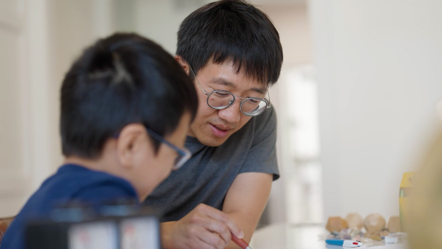 An adult and a child, both wearing glasses, sit closely together at a table. The adult is helping the child with an activity, possibly drawing or homework. The background is softly blurred.