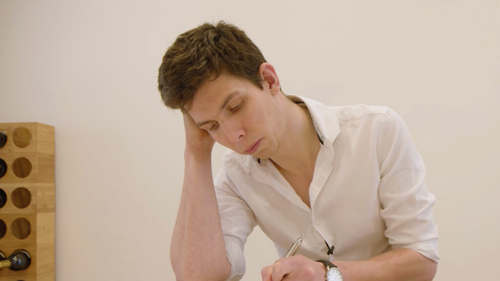 A man in a white shirt sits at a table, resting his head on one hand and writing with the other. He appears focused or thoughtful. A wooden wine rack is visible in the background.